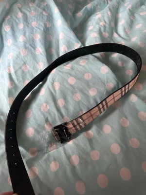 Beige och svart randigt bälte - Säljer ett stilrent bälte med klassiskt beige och svart randigt mönster. Bältet har en svart spänne och är tillverkat i skinn. Perfekt för att lyfta din outfit med en ikonisk detalj.