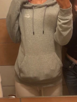 Grå hoodie från Nike med huva - Säljer en ljusgrå hoodie från Nike med klassisk logga på bröstet. Hoodien har en stor känguruficka framtill, justerbar dragsko i huvan och mjukt material som känns skönt mot huden. Perfekt för en avslappnad och sportig stil.