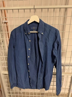 Blå slim fit skjorta från Ralph Lauren - Snygg blå skjorta från Ralph Lauren i modellen Slim Fit Indigo Oxford. Skjortan har klassisk krage med knappar, långa ärmar och diskret logga broderad på bröstet. Tillverkad i mjuk bomull för en bekväm känsla och stilren look.