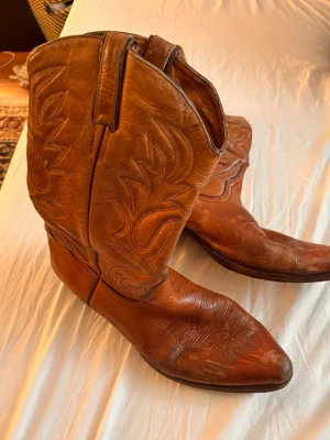 Bruna klassiska cowboy boots i läder - Säljer ett par bruna cowboy boots i äkta läder med snygga broderade detaljer längs skaftet och foten. Bootsens spetsiga tå och robusta klack ger en autentisk westernkänsla. Perfekta för dig som vill ha en tidlös och cool stil. Köpta för 2000kr!