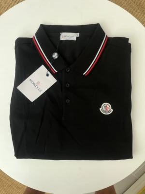 Svart Moncler pikétröja med logga - Klassisk svart pikétröja från Moncler med vit och röd rand på kragen. Tröjan har knappar framtill och Monclers ikoniska logga broderad på bröstet. Perfekt för en clean och stilren look.