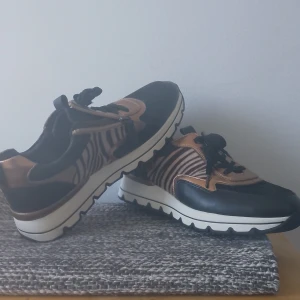 Zebramönstrade sneakers med metallicdetaljer - Snygga sneakers från Tamaris i svart och brunt med zebramönstrade sidor och metallic-kopparfärgade detaljer. Skorna har snörning, dragkedja på sidan och en chunky vit sula med svarta detaljer. Ovandelen är i skinn och syntet, perfekt för dig som gillar att sticka ut.