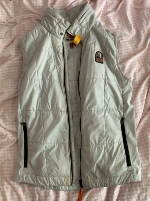 Ljusgrå dunväst från Parajumpers - Ljusgrå dunväst från Parajumpers med hög krage och två svarta dragkedjefickor. Västen har ett snyggt PJS Anchorage-märke på bröstet och en gul detalj vid halsen. Perfekt för lager-på-lager och coola höstdagar.