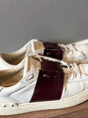 Valentino vita sneakers med vinröda detaljer - Snygga vita sneakers från Valentino med breda vinröda läderdetaljer över snörningen och klassiska nitar på hälen. Skorna har en chunky sula och är tillverkade i läder för en exklusiv känsla. Perfekta för dig som vill ha en stilren men ändå unik look.