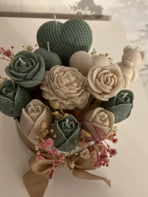 Dekorativt doftljus bukett i box - Unikt doftljus i rund vit box med beige rosett. Innehåller handgjutna ljus i form av blommor och hjärtan i grönt och vitt, samt små torkade blommor och en liten björndekorations ljus. Perfekt som present eller dekoration till rummet.