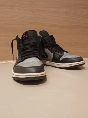 Nike Air Jordan 1 Mid svart/grå - Snygga Nike Air Jordan 1 Mid sneakers i svart och grått läder med svarta skosnören och vit sula. Klassisk Swoosh på sidan och röd Jumpman-logga på plösen samt rött Air Jordan-märke på ankeln. Perfekta för dig som gillar streetwear och ikonisk design.