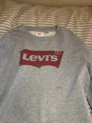 Grå sweatshirt från Levi's - Säljer en klassisk grå sweatshirt från Levi's med det ikoniska röda logotrycket på bröstet. Tröjan har rund halsringning och är tillverkad i mjukt bomullsmaterial. Perfekt för en avslappnad och stilren look.