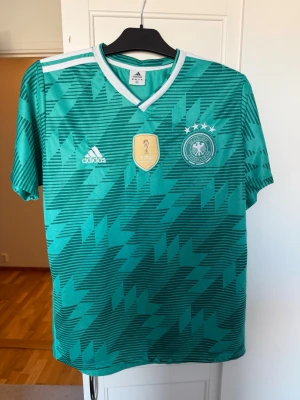 Grön fotbolls t-shirt Adidas Tyskland  - Säljer en grön t-shirt från Adidas med Tysklands landslagslogga och FIFA-märke på bröstet. T-shirten har ett grafiskt mönster, vita detaljer och korta ärmar. Perfekt för dig som gillar fotboll och vill ha en cool supportertröja.