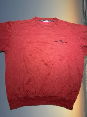 Röd crewneck från Sergio Tacchini - Röd crewneck-tröja från Sergio Tacchini med broderad logga på bröstet. Klassisk passform med rund halsringning och ribbade muddar vid ärmslut och nederkant. Perfekt för en avslappnad och stilren look.