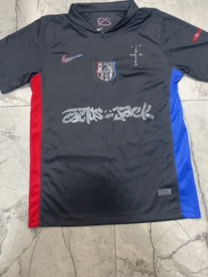 Nike x Cactus Jack FC Barcelona tröja - Svart kortärmad fotbollströja från Nike i samarbete med Cactus Jack och FC Barcelona. Tröjan har röda och blå paneler på sidorna, klubbmärke på bröstet och Cactus Jack-grafik framtill. Perfekt för dig som gillar unika sportkläder och streetwear.