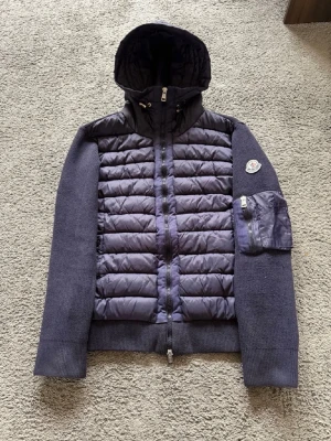 Mörkblå Moncler jacka med huva - Snygg mörkblå jacka från Moncler med quiltad front och huva. Jackan har stickade ärmar och rygg, samt en praktisk ficka med dragkedja på ena ärmen. Perfekt för dig som vill ha både stil och funktion. Den är i storlek m och har NFC samt qr kod som gör att du kan scanna och komma in på Monclers hemsida 