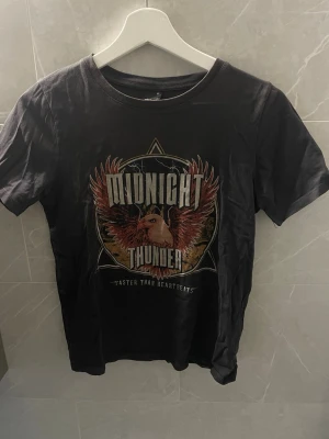 Svart t-shirt  - Cool svart t-shirt från Midnight Thunder med ett stort eagle print och texten 'Faster than Heartbeats' på framsidan. Klassisk rund hals och korta ärmar.