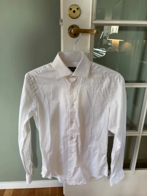Vit slim fit skjorta från Dressmann - Storlek XS men passar även S. Klassisk vit skjorta från Dressmann i slim fit-modell. Skjortan är tillverkad i bomull med stretch för extra komfort och har långa ärmar samt knäppning framtill. Perfekt för en clean och stilren look. 