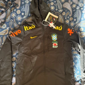 Brazil jacka - Säljer en svart Nike jacka med huva från Brasiliens landslag. Jackan har loggor och text i gult och orange på ärmarna, samt Brasil-märke på bröstet. Materialet är troligtvis polyester och jackan har dragkedja framtill och fickor. Perfekt för sport och träning.