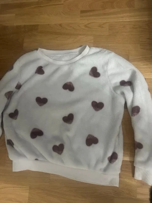 Mysigt loungewear-set med hjärtan - Supermjukt loungewear-set med vit tröja dekorerad med lila hjärtan och matchande lila byxor. Båda plaggen är i fluffigt material och har avslappnad passform – perfekt för hemmamys. Tröjan har rund hals och långa ärmar, byxorna har resår i midjan.