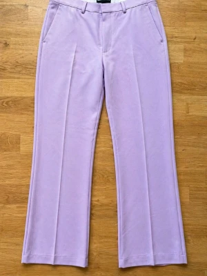 Rosa/lila utsvängda kostymbyxor ASOS - Storlek midja 32, längd 34. Säljer ett par ljuslila kostymbyxor från ASOS DESIGN med utsvängda ben och pressveck. Byxorna har fickor i sidorna och hällor för skärp. Perfekta för dig som vill sticka ut med färg och stil.