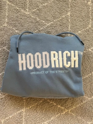 Blå HOODRICH hoodie med tryck - Snygg blå hoodie från HOODRICH med stort vitt broderat tryck på bröstet och texten 'PRODUCT OF THE STREETS'. Klassisk känguruficka framtill och justerbar huva med dragsko. Perfekt för en streetwear-look.