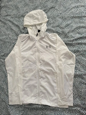 Under Armour vindjacka vit - Snygg vit vindjacka från Under Armour med huva och hel dragkedja. Jackan har logga på bröstet och stor text på ryggen. Tillverkad i lätt och tunt material som passar perfekt för blåsiga dagar.