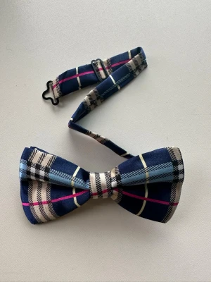 Helt NY blå rutig fluga - Snygg fluga med rutigt mönster i blått, beige, vitt och rosa. Flugan har ett justerbart band med metallspänne och är tillverkad i ett mjukt satintyg. Perfekt för att ge din outfit en cool touch.