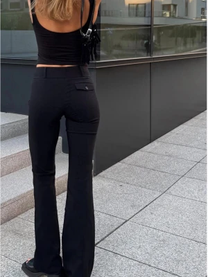 Svarta kostymbyxor med utsvängda ben - Snygga svarta kostymbyxor med utsvängda ben och hög midja. Byxorna har en klassisk design med fickor bak och knäppning framtill. Perfekta för dig som vill ha en stilren och trendig look. Materialet är slätt och ger en elegant känsla.