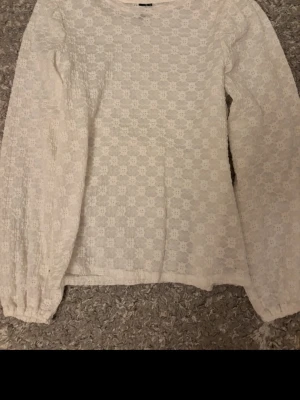 Vit spetsblus från Vero Moda - Söt vit spetsblus från Vero Moda med blommigt mönster i spets och rund halsringning. Blusen har långa ärmar med resår vid ärmslut och är tillverkad i ett tunt, genombrutet material som ger en romantisk vibe.