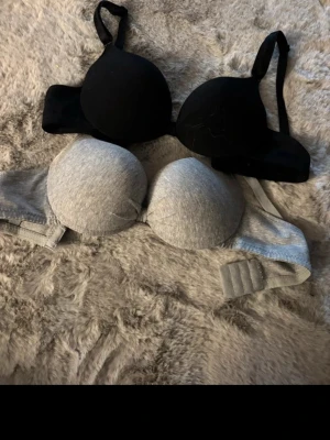 2-pack bhar svart och grå - Säljer två bh:ar med vadderade kupor, en i svart och en i ljusgrått. Båda har justerbara axelband och hakknäppning bak. Materialet är mjukt och stretchigt, perfekt för en bekväm känsla hela dagen. Inga synliga märken på bh:arna.