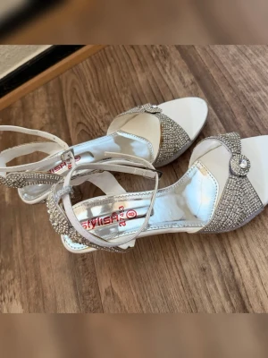 Silvriga klackar med strass och remmar - Säljer ett par glittriga klackar i silver med öppen tå, dekorerade med strass och en liten brosch framtill. Skorna har smala remmar runt ankeln och en klack täckt av stenar för extra bling. Klackhöjd cirka 6cm. Perfekta för dig som vill sticka ut och glänsa. Köptes utomlands och har använts ett par gånger. Skriv gärna om du har frågor eller vill se ytterligare bilder. Det står storlek 8 på klacken och passar en med storlek 37/38. 