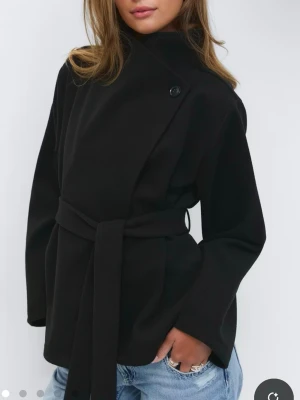 Svart kappa från Gina  - Stilren svart kappa med omlottknäppning och knytband i midjan. Hög krage med knappdetalj och raka ärmar. Perfekt för en trendig och minimalistisk look. Passar till jeans eller dressat. Materialet ser ut att vara ullblandning.