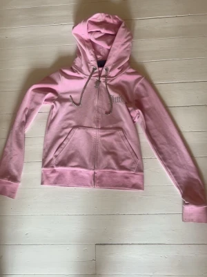Rosa Juicy Couture hoodie med dragkedja - Säljer en ljusrosa hoodie från Juicy Couture med dragkedja framtill, huva med glittriga dragskor och fickor på magen. Märkets logga i strass på bröstet ger en extra touch. Perfekt för en chill och trendig look.