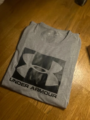 Grå Under Armour t-shirt - Snygg grå t-shirt från Under Armour med stort svart logotryck på bröstet. Tillverkad i mjukt och lätt material med rund halsringning och relaxed fit. Perfekt för träning eller chill dagar.