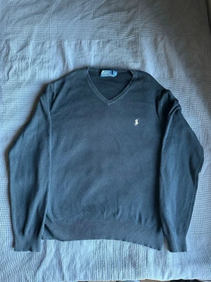 Mörkblå v-ringad tröja Polo Ralph Lauren - Klassisk svart v-ringad tröja från Polo Ralph Lauren med den ikoniska broderade loggan på bröstet. Tröjan har långa ärmar och ribbade muddar vid ärmslut och nederkant. Perfekt för en stilren och avslappnad look.