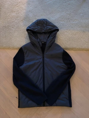 Mörkblå J.Lindeberg jacka - Snygg mörkblå jacka med vadderad framsida och huva. Ärmar och rygg är ribbstickade för extra komfort och stil. Jackan har dragkedja framtill och en sportig look som passar perfekt till vardags. Enkel och modern design.