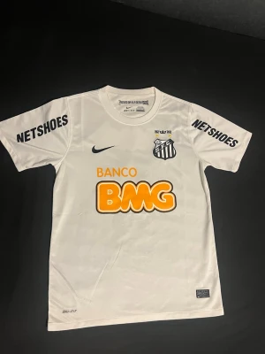 Neymar Jr #11 Santos tröja - Vit fotbollströja från Nike med Santos FC klubbmärke på bröstet. Tröjan har tryck från sponsorer som BMG och Netshoes, samt Dri-FIT material som håller dig torr under träning. Rund halsringning och korta ärmar. Perfekt för dig som gillar brasiliansk fotboll.