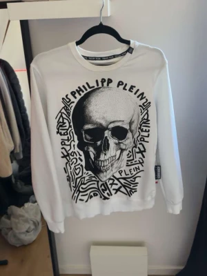 Vit sweatshirt från Philipp Plein - Cool vit sweatshirt från Philipp Plein med svart grafiskt tryck och logotyp på framsidan. Rund halsringning och ribbade muddar vid ärmslut och nederkant. Perfekt för dig som gillar streetstyle och unika prints.