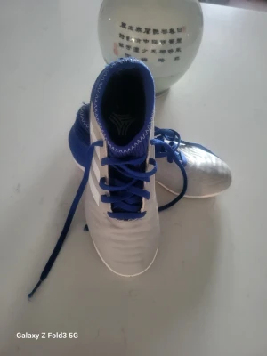 Adidas blåvita fotbollsskor - Snygga fotbollsskor från Adidas med vit ovandel och blå detaljer, inklusive snörning och häl. Skorna har klassiska tre vita ränder på sidan och är tillverkade i syntetmaterial som ger bra komfort och grepp på planen.