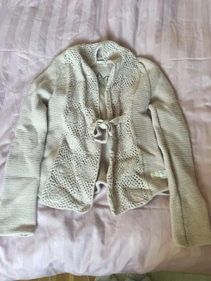 Rosa stickad kofta med knytband - Söt stickad kofta från Odd Molly i en gammalrosa färg med dekorativa virkade detaljer och blommönster framtill. Koftan har ett knytband i midjan och långa ärmar. Även en broderad liten ängel på ryggen 🥰