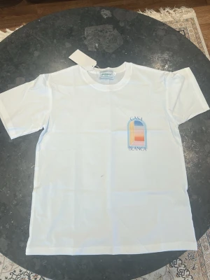 Vit Casa Blanca t-shirt med tryck - Vit t-shirt från Casa Blanca med färgglatt grafiskt tryck på bröstet och ryggen. Trycket har en båge i blått, orange och rosa med texten 'Casa Blanca'. Klassisk passform och rund hals, perfekt för en chill look.