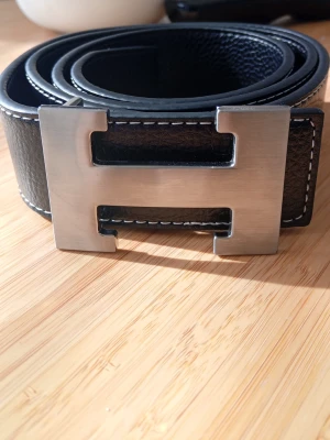 Svart skärp med stor H-spänne - Snyggt svart skärp med ett stort, silverfärgat H-spänne. Skärpet har vit kontrastsöm längs kanterna och är gjort i skinn med en stilren och klassisk look. Perfekt accessoar för att lyfta vilken outfit som helst. DM för mer info/ diskutera pris!! 120 cm långt