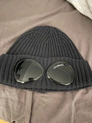 Svart mössa från C.P. Company - Säljer en svart ribbstickad mössa från C.P. Company med de ikoniska inbyggda goggles framtill. Mössan är tillverkad i ull och har en uppvikt kant. Perfekt för dig som vill sticka ut med en snygg look. Den har mycket bra skick och använd max 5 gånger. 