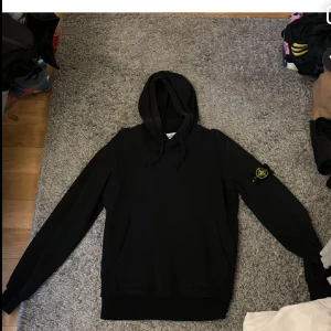 Svart hoodie från Stone Island - Svart hoodie från Stone Island med klassisk huva och logotyp-patch på vänster ärm. Tröjan har långa ärmar, ribbade muddar och känguruficka framtill. Perfekt för en avslappnad och stilren look.