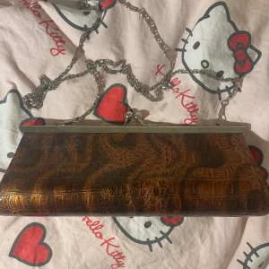 Brun kuvertväska med krokopräglat mönster - Så fin och elegant clutch. Kjedjan går att ta av så man kan ha den både runt axeln eller i handen. Köpt second hand men aldrig använd av mig