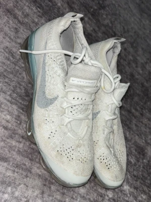 Nike Air Vapormax vita sneakers - Säljer ett par Nike Air Vapormax sneakers i vitt med grått swoosh på sidan. Skorna har stickad ovandel i mesh och genomskinlig bubbelsula som ger en futuristisk look. Perfekta för dig som gillar sportig stil och komfort.