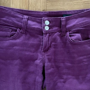 Lila jeans - Snygga lila manchesterbyxor med raka ben och dubbla vita knappar framtill. Byxorna har klassiska fickor både fram och bak, samt en bakficka med knappstängning. Perfekt för dig som vill sticka ut med färg och struktur.