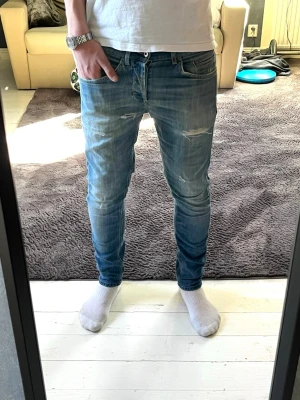 Blå dondup jeans - Säljer ett par blåa dondup jeans med slitningar på låren och knäna. Jeansen har klassiska fem fickor och är tillverkade i denim med stretch för skön passform. Det är modellen George och storlek 32. En liten lagning också som ni ser på bild 7. Hör av er om ni har några frågor!☺️