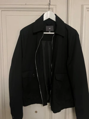 Svart vindjacka från H&M - Stilren svart vindjacka från H&M med rak passform, dragkedja framtill och två stora fickor med lock och knapp. Jackan har klassisk krage och elastisk nederkant för en snygg siluett. Perfekt för blåsiga dagar och enkel att matcha med olika outfits.