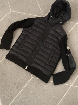 Svart pufferjacka från Moncler - Snygg svart pufferjacka från Moncler med quiltad front och huva. Jackan har stickade ärmar och muddar, dragkedja framtill och två fickor med dragkedja. Moncler-logga på ärmen och coolt innerfoder med serietidningsmotiv.