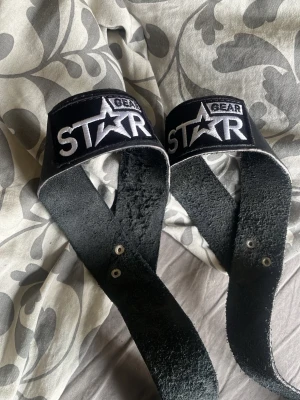 Svarta straps från Star Gear - Säljer ett par svarta straps från Star Gear. Remmarna är tillverkade i slitstarkt läder med broderad logga i vitt och svart. Perfekta för styrketräning och gym, ger extra grepp vid tunga lyft.