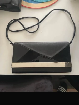 Svart kuvertväska med metallband - Stilren svart kuvertväska med lång axelrem och snyggt geometriskt lock. Väskan har en blank finish och ett dekorativt silverfärgat metallband på framsidan. Perfekt för att lyfta din outfit med en modern touch. Materialet är konstläder.