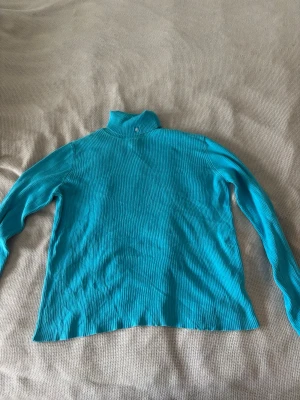 Turkos ribbstickad polotröja RL - Snygg turkos ribbstickad polotröja från Ralph Lauren med diskret RL-brodyr på kragen. Tröjan har lång ärm och är tillverkad i ett mjukt, stretchigt material som sitter skönt på kroppen. Perfekt för lager på lager och en färgklick i garderoben.