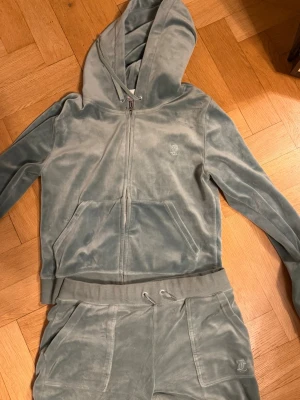 Grå velour loungewear-set Juicy Couture - Mysigt loungewear-set från Juicy Couture i grå velour. Setet består av en zip-hoodie med fickor och logga på bröstet samt matchande byxor med dragsko och logga på höften. Perfekt för chill dagar hemma.